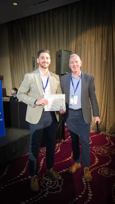Two men in business attire stand on a patterned carpeted floor, one holding a certificate, both wearing conference badges and lanyards.