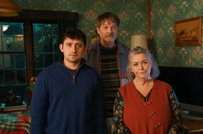 Craig Roberts, Mark Heap and Dawn French look to camera dressed in warm home clothing in a cosy looking house