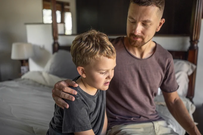 A man comforting young boy while sitting on a bed.
