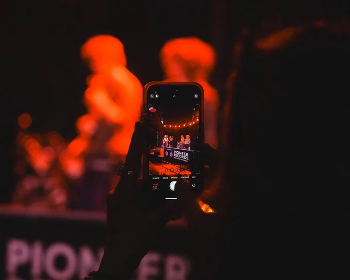 A person records a live concert on their smartphone, capturing a band performing on stage, illuminated by red stage lighting, with the Pioneer logo visible on DJ equipment. Photo by Jordan Kullen '25