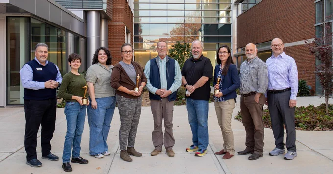 Group of nine professionals holding awards, standing in front of a modern academic building.
