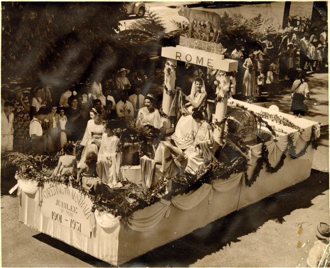 A sepia photo of a float designed to evoke Ancient Rome in a parade in Cairns. In the float sits young ladies dressed as ancient Roman muses. The banner reads "Greetings to Australia, Jubilee, 9th May, 1901-1951.