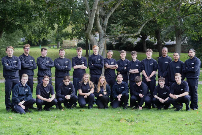 A group of 22 UK Power Networks foundation apprentices pose together for a group photo. They are standing in a park outside.
