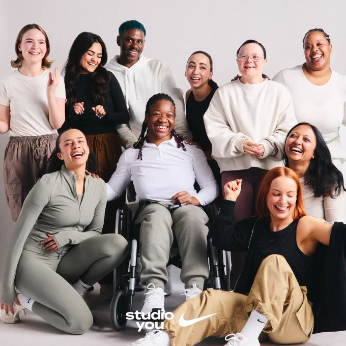 A diverse group of individuals posing together, smiling in a studio setting, featuring a "Nike" and "Studio You" logo at the bottom. Some are standing while others are seated, including one person in a wheelchair. They wear casual sportswear.