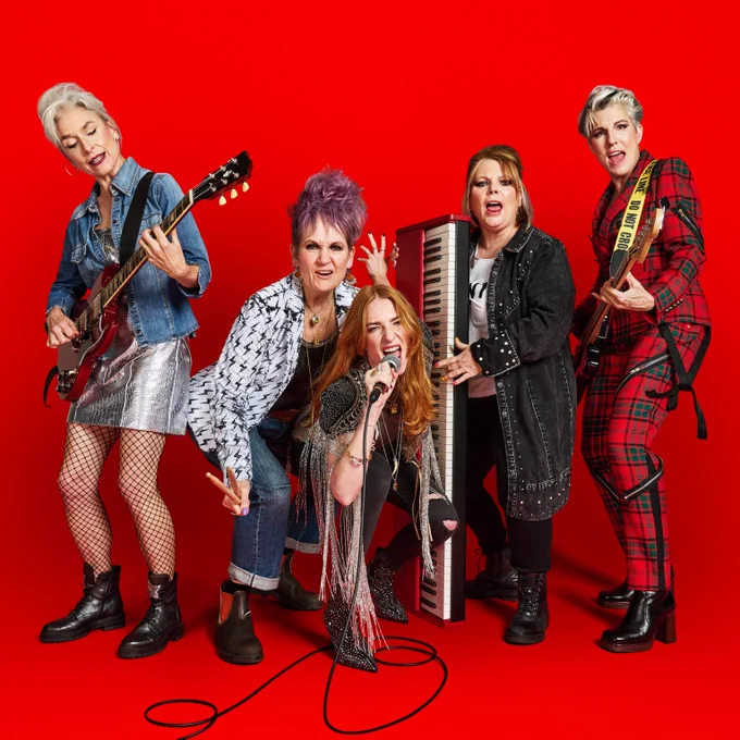 Five women holding various musical instruments, dressed in punk and rock clothing. They pose together in a group against a red background. 