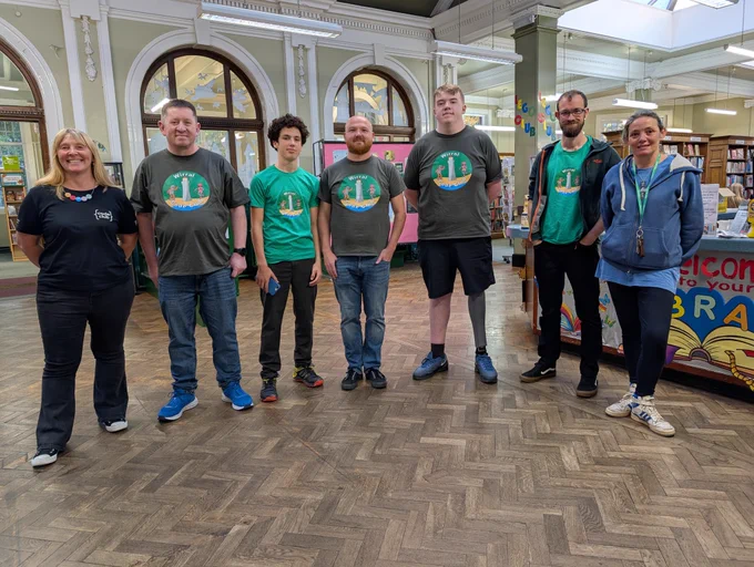 Seven people standing in a library with wooden floors, some wearing matching green t-shirts featuring a circular logo with trees and a lighthouse.