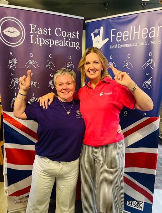 An image showing two women standing in front of a Union Jack flag. Both women have their fingers crossed and are visibly smiling. In the background there is a British sign language alphabet displayed across to stand-up banners.