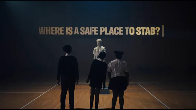 Students stand in empty gym hall looking at a life size human figure, with the question: 'Where is a safe place to stab?' projected on the wall.