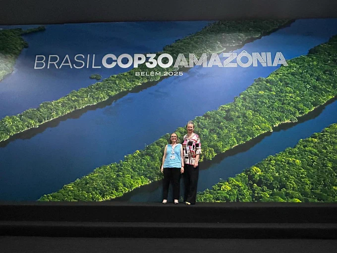 Elizabeth Kujawinski and Sonya Dyhrman stand in front of a backdrop of the Amazon River, underneath a sign that states Brasil COP30 Amazonia, Belem 2025. Photo by Elizabeth Kujawinski, WHOI. 