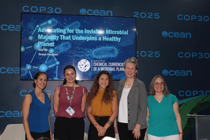 The panel participants (left to right: Marcia Macedo, Pearl Mitchell, Mariana Rocha de Souza, Sonya Dyhrman, and Elizabeth Kujawinski) stand in front of a monitor displaying the name of the panel, Advocating for the Invisible Microbial Majority that underpins a healthy planet, in the Ocean Pavilion at COP30. Photo by Danielle Fino, WHOI.