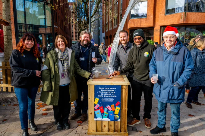 A group of people standing outdoors at a festive market beside a wooden donation station with a sign encouraging the donation of empty glasses to support charity. The scene includes some individuals wearing seasonal accessories like a Santa hat.