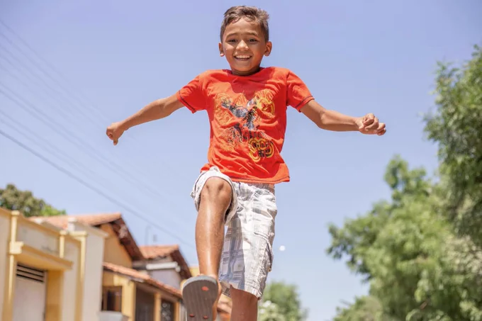 A child in an orange t-shirt and white shorts playing hopscotch on a sunny day.