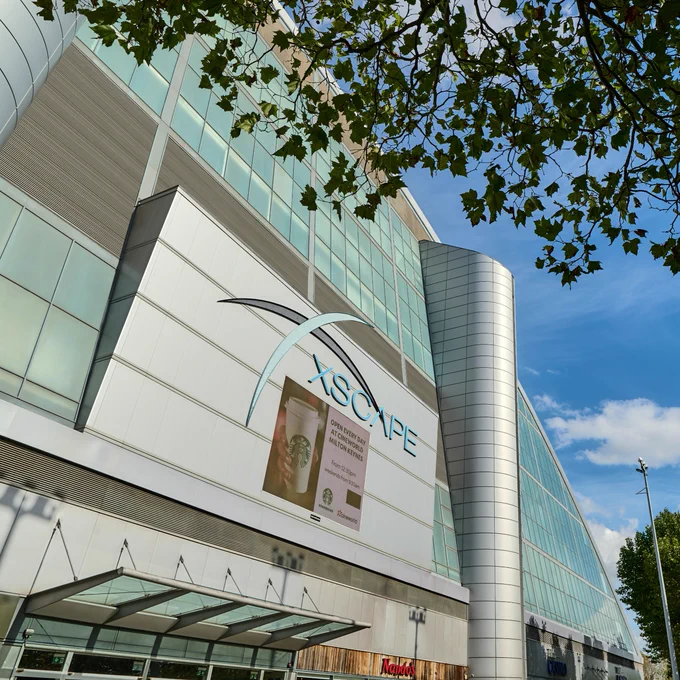 Exterior view of the Xscape Milton Keynes building on a sunny day, adorned with banners and surrounded by green foliage.