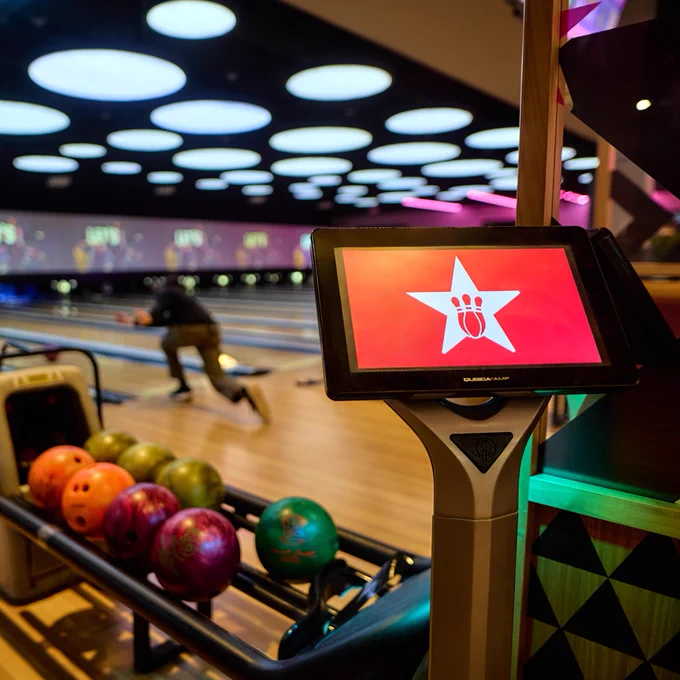 A Hollywood Bowl bowling scoring monitor with colourful bowling balls in the foreground and bowlers in action in the background under blue-lit ceiling.