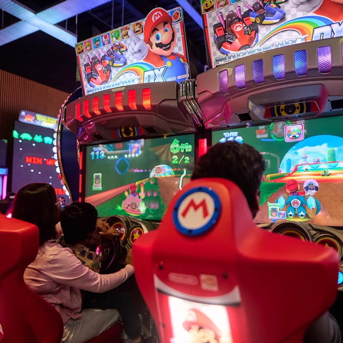 Individuals playing a Mario Kart arcade game in a vibrant arcade setting.