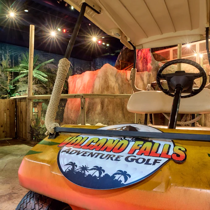 An adventure golf cart labeled "Volcano Falls Adventure Golf" parked in a themed indoor setting with tropical and volcanic rock features.