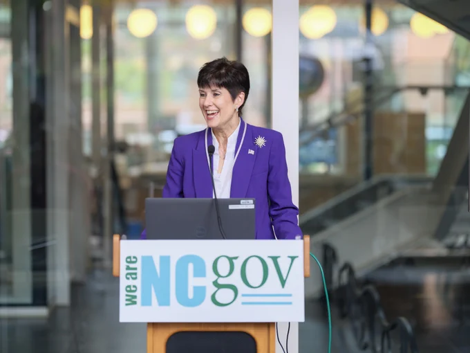 Secretary Piccone stands at a podium in a purple blazer smiling during a presentation in front of a glass backdrop with a sign reading "We Are NC Gov."