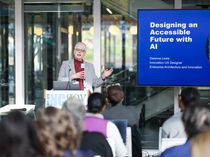 Person presenting at a conference with a slide titled "Designing an Accessible Future with AI" Audience is attentively listening in a modern conference room with glass walls.