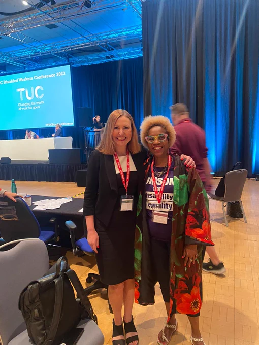 Vicky (left) stood smiling with a UNISON disability group member (right) in front of a conference hall with seats and a bright blue screen showing 'TUC'.
