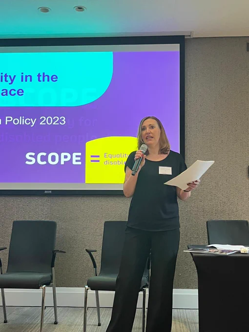 Vicky stands talking on a stage into a microphone with paper in her hands. Behind her is a purple screen displaying SCOPE's logo. 