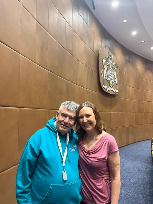 Vicky (right) stood smiling beside a Lewisham Speaking Up member (left). They are stood in the Lewisham Council Chamber in the Civic Centre. Behind them, is a brown curved wall displaying the Borough of Lewisham coat of arms.  