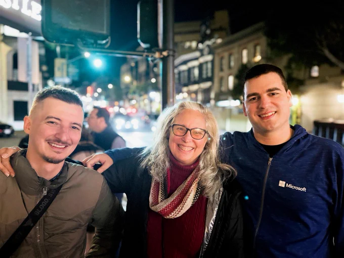 Three people standing close together outdoors at night on a city street, with bright lights and blurred traffic in the background. One person is wearing a dark jacket with a crossbody strap, another has a scarf draped over a red top, and the third is in a blue Microsoft-branded hoodie.