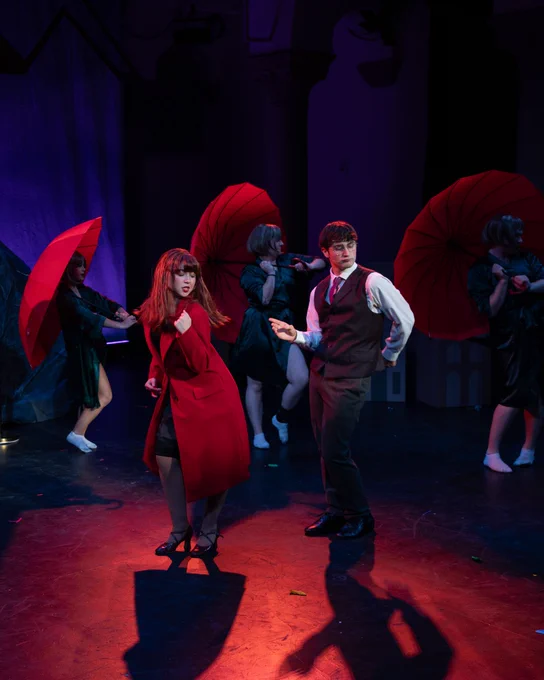 Performers with red umbrellas on stage in a theatrical production, featuring dynamic poses and expressive costumes.