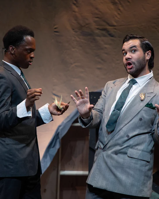 Two actors are performing a dramatic scene on stage, one wearing a badge on their suit. They are engaged in a lively conversation, using expressive gestures.