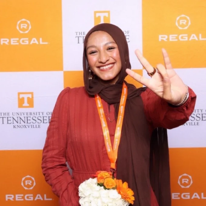 Smiling individual in a hijab holding a bouquet and making a 'Vols' sign, standing in front of a banner with the University of Tennessee, Knoxville, and Regal logos.