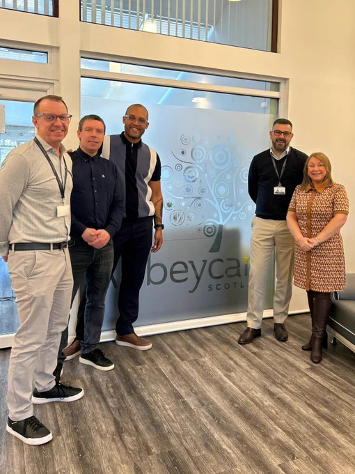 Five people stand together smiling inside a bright, modern building with a frosted glass panel featuring the Abbeycare logo behind them. They appear to be posing for a group photo after a meeting or visit.