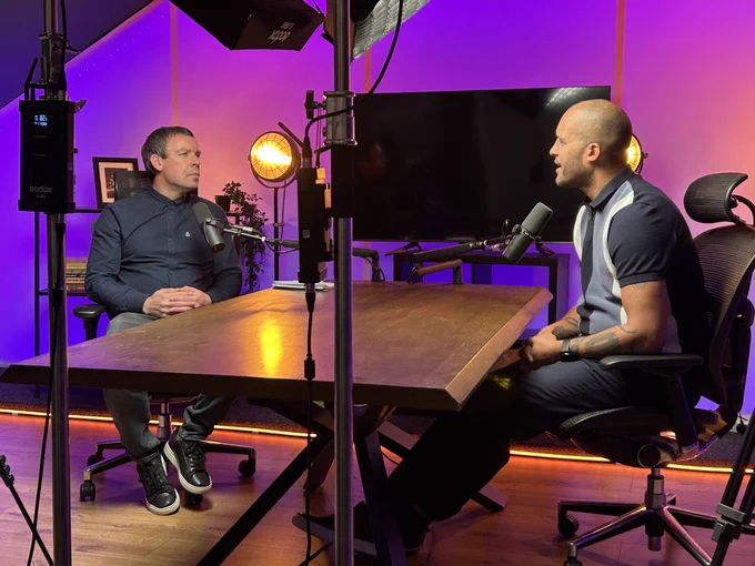 Side-angle view of two men recording a podcast in a colourful, professionally lit studio. They sit facing each other across a large wooden desk, each speaking into mounted microphones. Lighting equipment is visible around them.