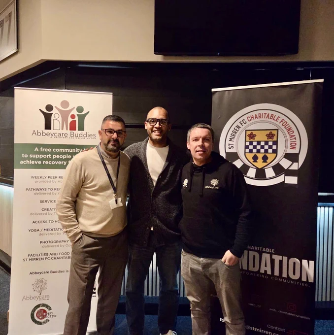 Three men stand together smiling in front of two banners—one for a recovery support programme and one for a football club charitable foundation. They are posing indoors in a casual event setting.