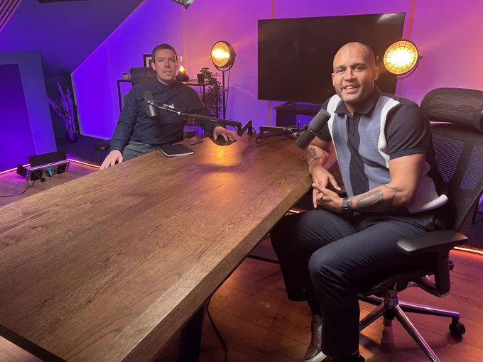 Two men sit at opposite sides of a wooden podcast table in a studio with purple and pink lighting. Each has a microphone set up in front of them. One man sits on the left with a notebook on the table, while the other sits on the right, smiling with folded hands. Studio lights, plants, and a TV screen are in the background.