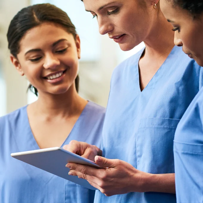 A photograph showing three healthcare workers in blue scrubs, reviewing some information on a tablet.