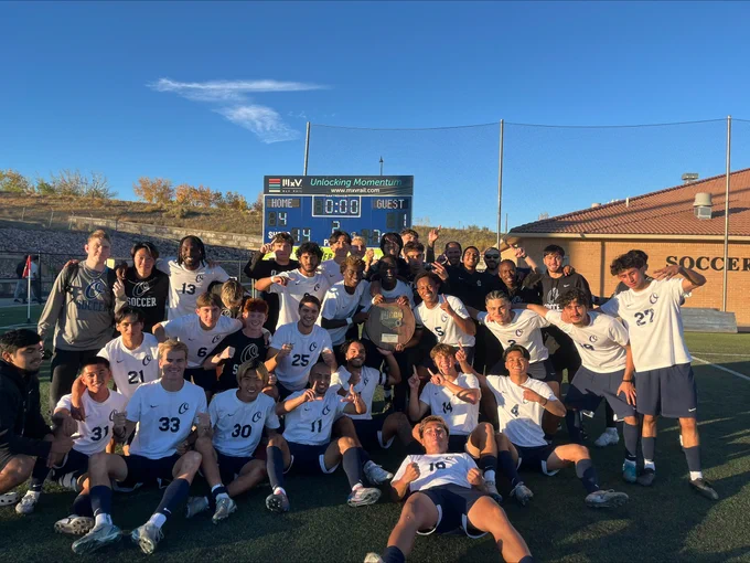 A large group of Otero College men’s soccer players and coaches pose together on the field in front of a scoreboard, smiling and holding up number-one gestures after a championship win.