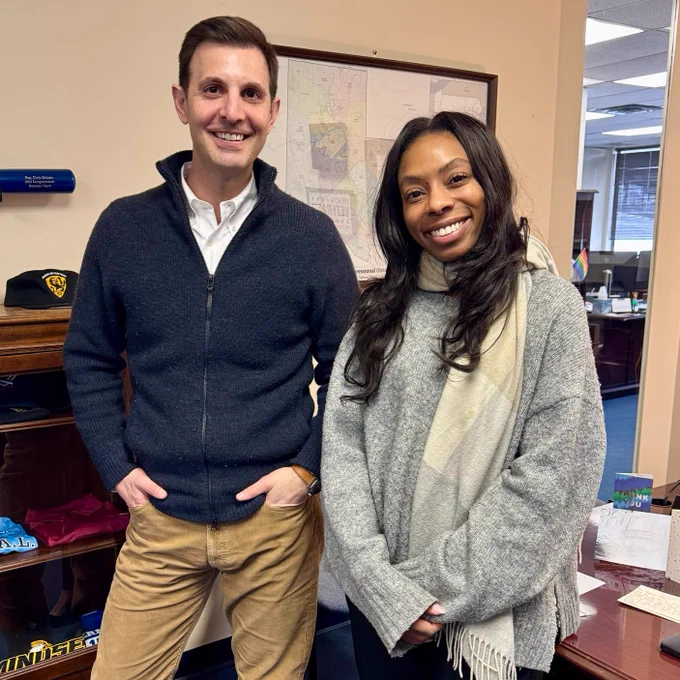 Two people stand side by side in an office. On the left is a man wearing a dark zip-up sweater, a white shirt, tan pants, and brown shoes. On the right is a woman wearing a loose gray sweater, a light scarf, black pants, and black heels. Both are smiling. A framed district map hangs behind them, and office furniture and display shelves are visible around them. The man is Congressman Chris Deluzio, and the woman is State Representative Lindsay Powell.