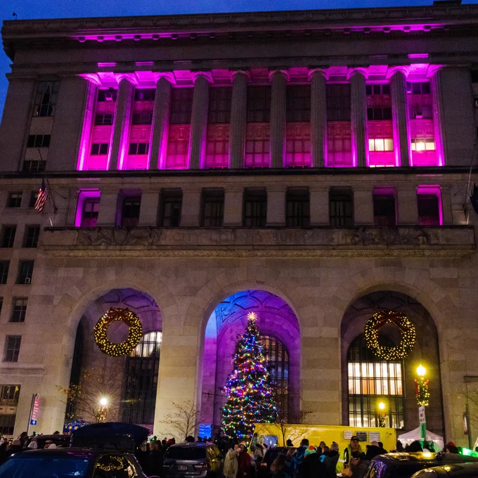 The City County Building in Downtown Pittsburgh is illuminated with pink lighting and decorated with large wreaths. A Christmas tree adorned with lights stands in front, surrounded by people.