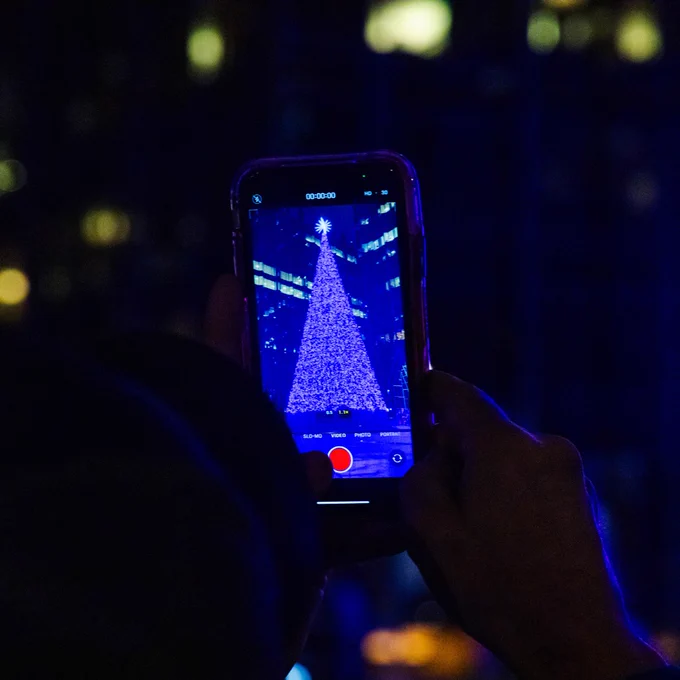 A person captures a photo of a Christmas tree illuminated at night on their smartphone screen.