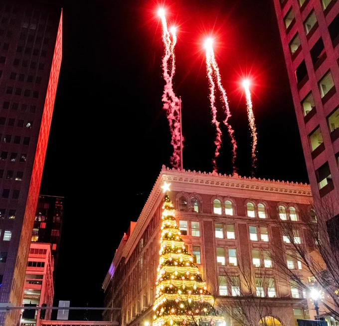 Fireworks burst above a large, illuminated Christmas tree on a building in Downtown Pittsburgh during a nighttime celebration. The surrounding buildings are dimly lit, enhancing the festive atmosphere.