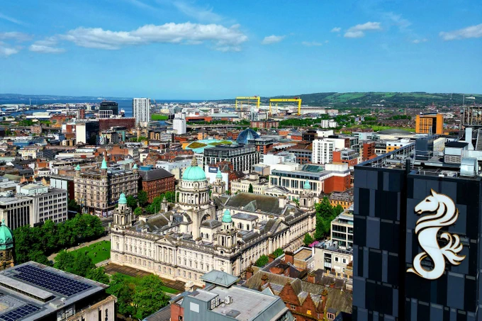Aerial view of Belfast city centre featuring Grand Central hotel and City Hall against blue sky.