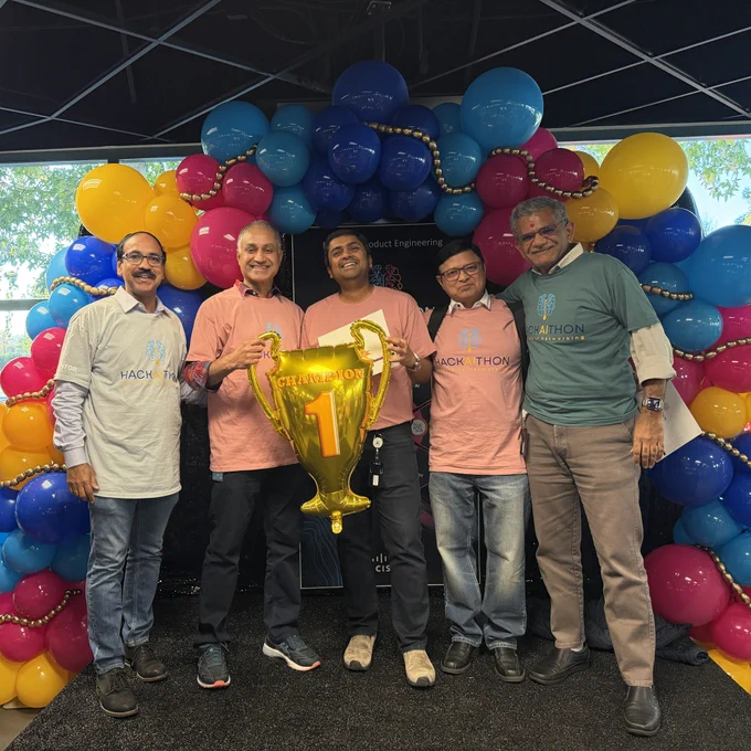 Five men stand on a stage in front of a colorful balloon arch, holding a large golden trophy shaped like the number '1'. Some wear pink "HACKATHON" t-shirts, and others wear light blue "HACKATHON" t-shirts. A banner behind them mentions "Product Engineering".
