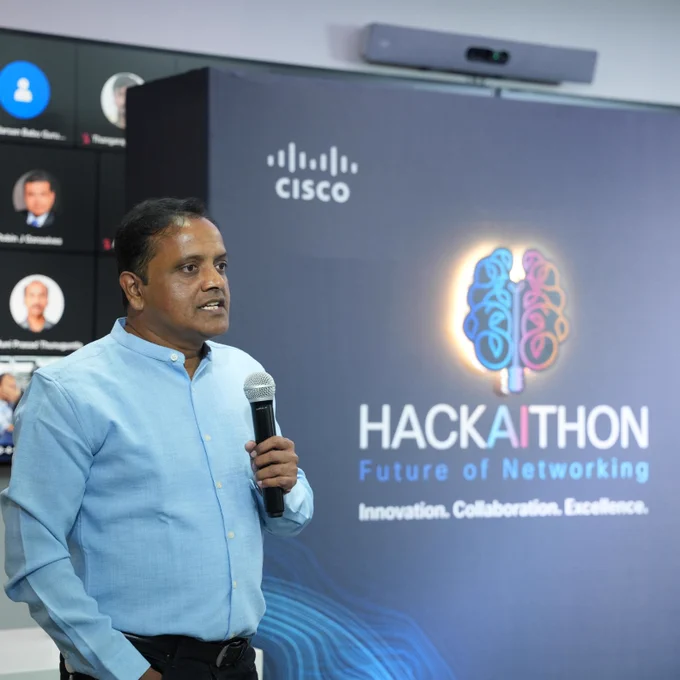 A man giving a presentation in front of a group at the hackathon event with a big hackathon blue sign behind him. 