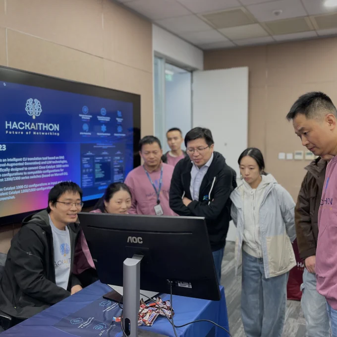 A group of hackathon participants stand around a desk, watching two teammates work at a computer monitor. A large screen on the wall displays “Hackaithon: Future of Networking” with technical details. The group appears focused and engaged as they review the project together.
