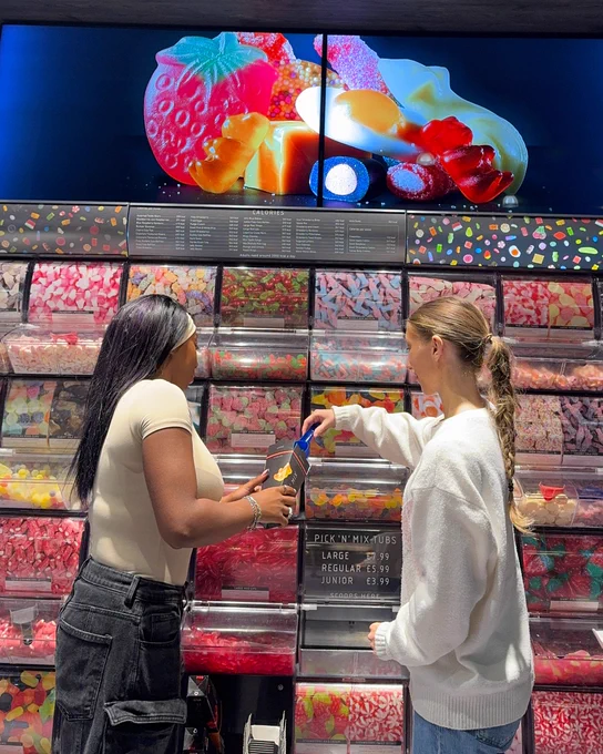 Two people selecting pick n mix from a large self-serve display in a brightly coloured store.