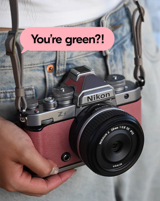 A close-up of a person holding a Nikon Z f Silver camera in Mauve Pink against their denim jeans. A pink speech bubble above the camera reads, “You’re green?!”