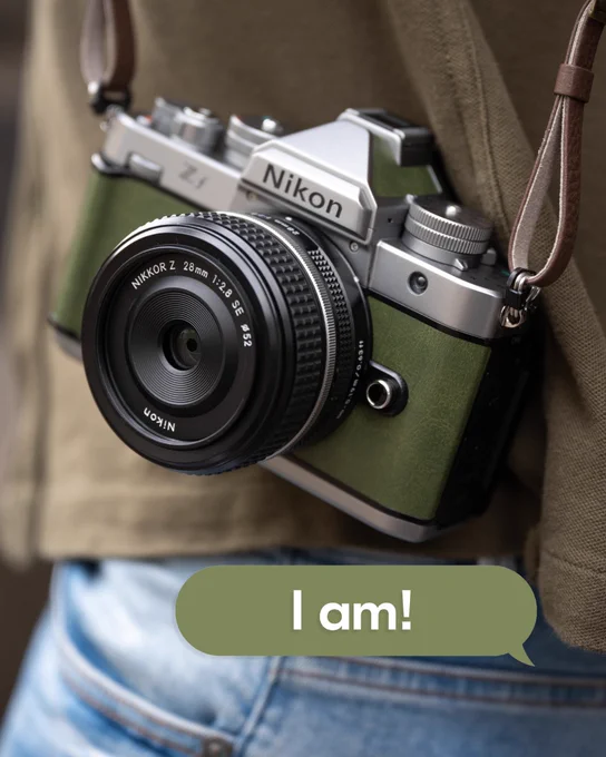A close-up of a green Nikon Z f Silver camera in Moss Green hanging against someone’s green shirt and denim jeans. A green speech bubble near the bottom reads, “I am!”