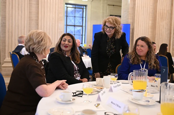 Four women are gathered around a circular table in a large, well-lit room with tall stone columns. Three women are seated, and one stands behind them, smiling. There are drinks, cups, and papers with names written on them on the table.