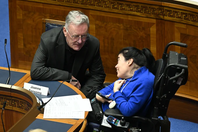 A close-up of a man in a dark suit leaning down to speak with a woman in a blue hoodie who is seated in a wheelchair. They are looking at each other and smiling. A name plaque for "Joanne Sansome" is visible on the desk in front of them, along with some papers.