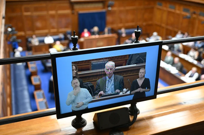 A close-up view of a monitor displaying a video feed of a man speaking from behind a desk, flanked by two women. Below the speaker, there are split screens showing two sign language interpreters, one male and one female. The image is taken from an elevated position, with the main chamber in the blurred background.