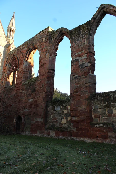 The images shows the red stone of the side wall of the Guesten in the early morning light on Friday. There are four windows in the images two very large and two half their size set into the stone. Behind the wall in the background can be made out a pinnacle in pale brown stone of the Cathedral. In the foreground is the lawn with frost on it. 